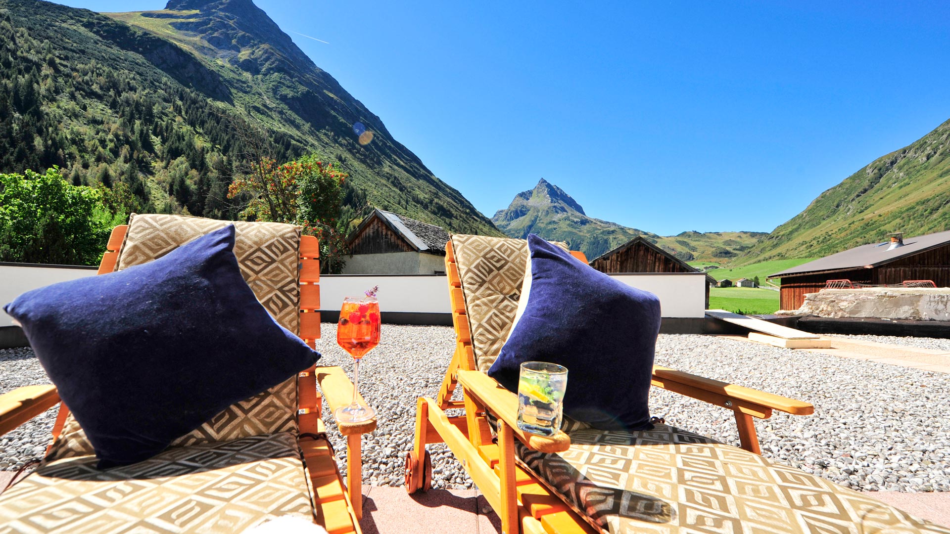Zwei Sonnenliegen mit Kissen und Cocktails auf Terrasse mit Blick auf grüne Berge – Sommeridylle pur.